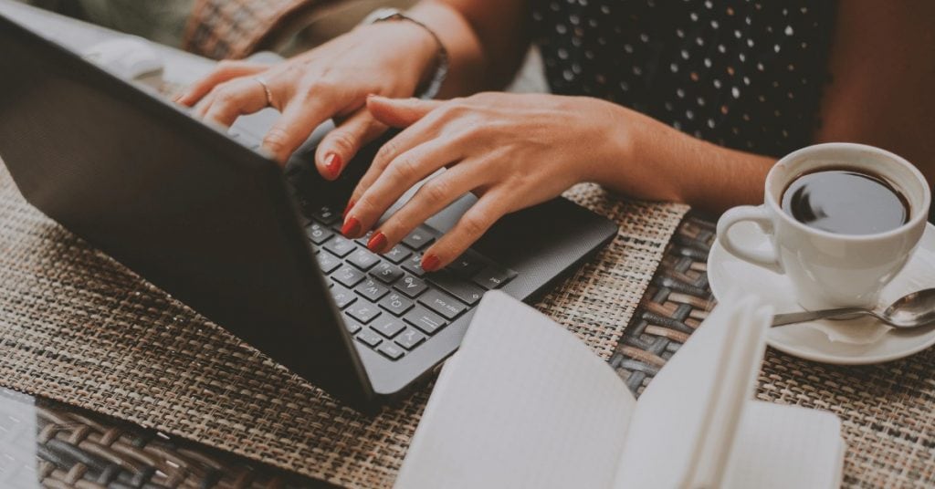 woman with red nails typing on a laptop while drinking coffee