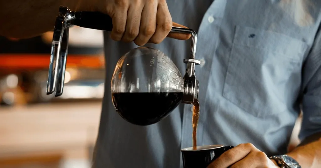 person pouring coffee from a siphon pot into a mug