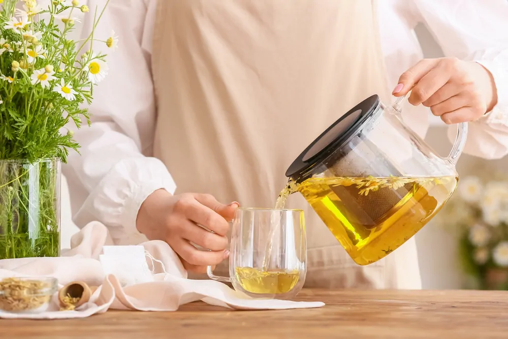 woman pouring tasty chamomile tea from teapot into cup
